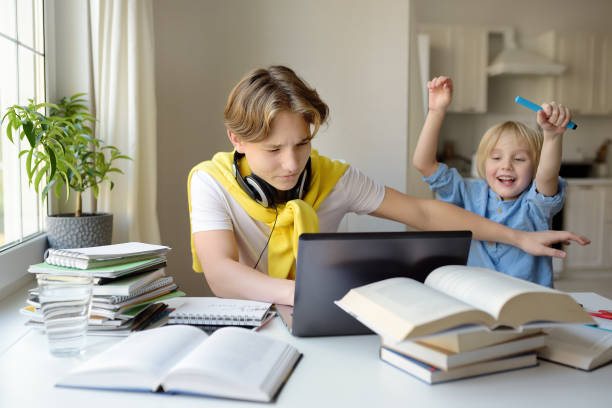 Teenager boy study at home and his younger brother screams and interrupts him. Online education and distance learning for high school students. School boy doing his homework Teenager boy study at home and his younger brother screams and interrupts him. Online education and distance learning for high school students. School boy doing his homework using gadgets. Distracted stock pictures, royalty-free photos & images