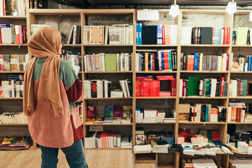 View from behind of muslim female student with hijab standing in library and looking on bookshelves