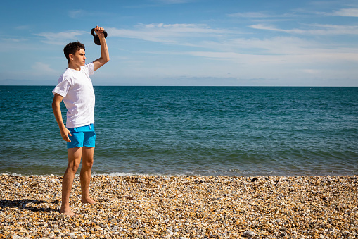 A 18 year old Caucasian teenage boy on a beach exercising with a kettlebell weight