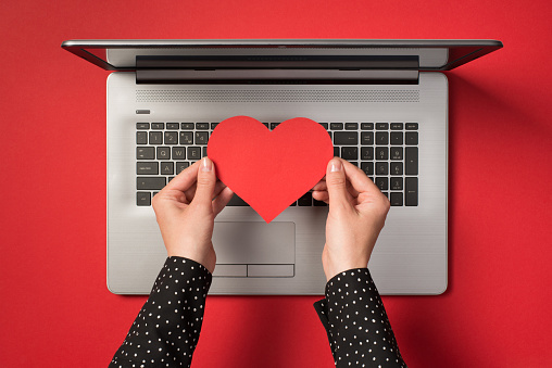 above photo of grey laptop and hands holding a red paper heart isolated on the red backdrop