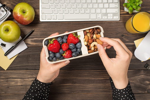 First person top view photo of woman's hands holding lunchbox with healthy meal nuts and berries over apples glass of juice flowerpot stationery keyboard mouse on isolated dark wooden table background