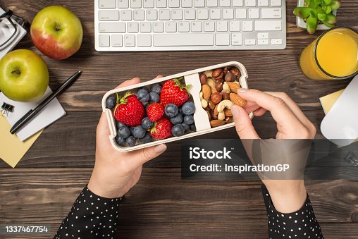 istock First person top view photo of woman's hands holding lunchbox with healthy meal nuts and berries over apples glass of juice flowerpot stationery keyboard mouse on isolated dark wooden table background 1337467754