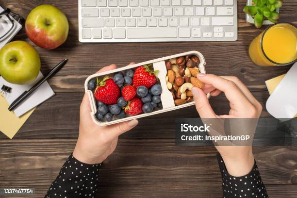 First Person Top View Photo Of Womans Hands Holding Lunchbox With Healthy Meal Nuts And Berries Over Apples Glass Of Juice Flowerpot Stationery Keyboard Mouse On Isolated Dark Wooden Table Background Stock Photo - Download Image Now