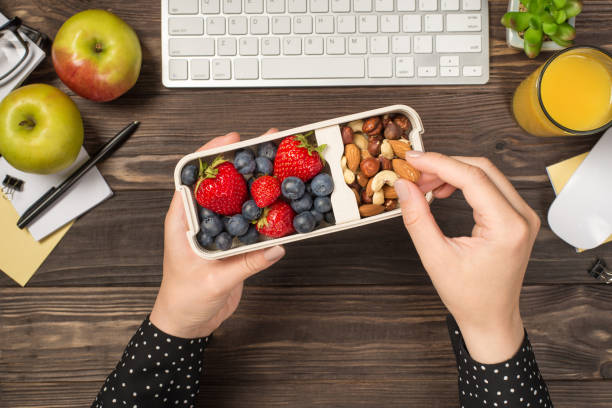 first person top view photo of woman's hands holding lunchbox with healthy meal nuts and berries over apples glass of juice flowerpot stationery keyboard mouse on isolated dark wooden table background - healthy food imagens e fotografias de stock