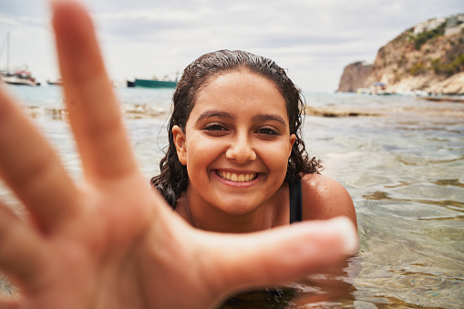 Young cheerful spanish woman approaching camera on natural pool in beach
