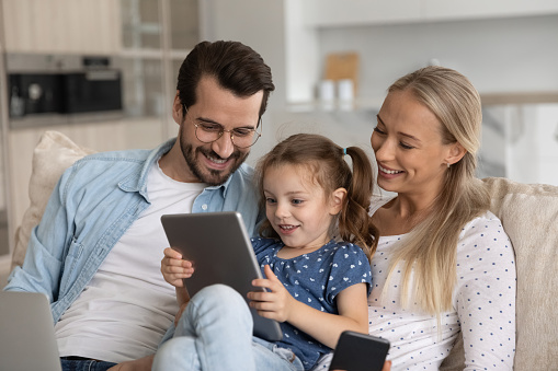 Happy excited gen Z little kid with tablet and parents with gadgets relaxing on couch, playing online game, looking at screen, smiling, laughing. Family using devices, making video call from home