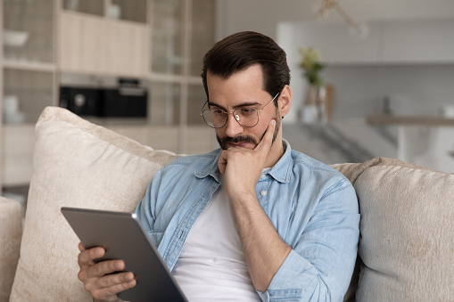 Focused millennial man wearing glasses, using tablet, reading email message with thoughtful face, thinking over text on screen, feeling doubt, concerned. Adult student watching learning webinar