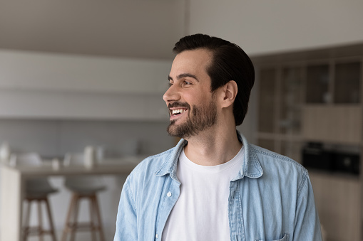 Happy millennial 30s man in casual home head shot portrait. Cheerful guy, homeowner, tenant posing in apartment, looking away with interest, laughing, smiling at positive thoughts, thinking of future