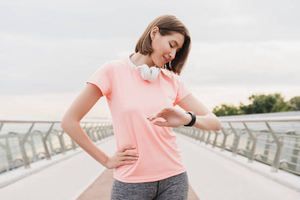 jovem atleta magra esportista verificando o tempo em seu relógio de pulso em roupas de ginástica se preparando para correr em uma ponte da cidade na área urbana. conceito de estilo de vida ativo - distance running audio - fotografias e filmes do acervo