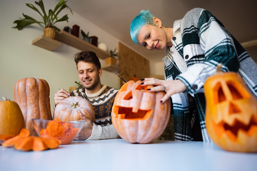 Young cheerful Caucasian couple preparing pumpkins for Halloween at home.
