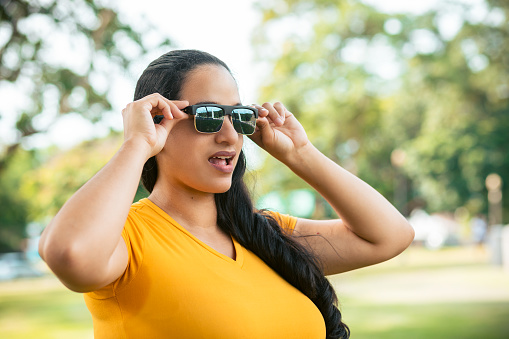 Young latin woman touching her sunglasses in the park amazed