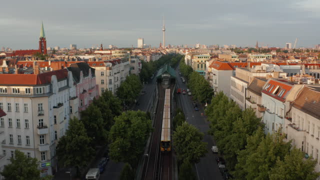 Forwards tracking scan train driving in wide street on large city. Roofs of buildings lit by bright morning sun. Fly towards Fernsehturm TV tower. Berlin, Germany