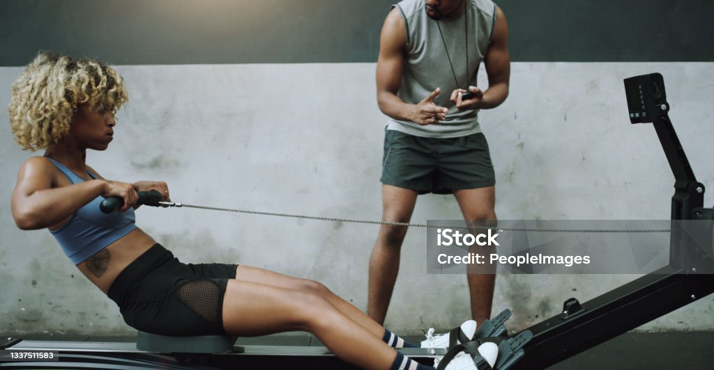 Shot of a young woman exercising on a cable row machine with a fitness trainer in a gym Beat the clock, become the best Exercising Stock Photo