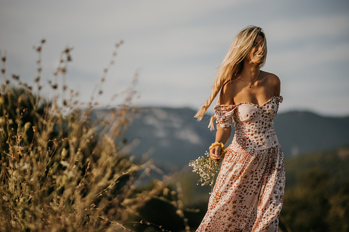 Young woman in dress holding bouquet in hand and walking in nature on sunny day