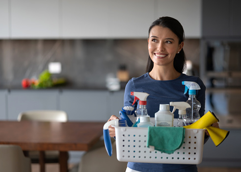 Happy Latin American woman at home holding a basket of cleaning products while doing the chores - domestic life concepts