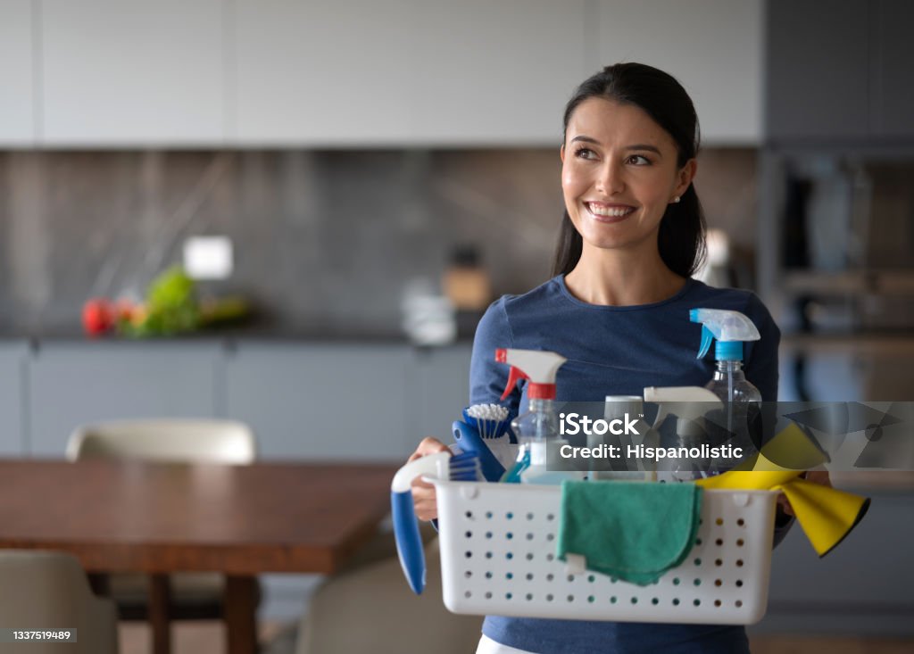 Happy woman at home holding a basket of cleaning products Happy Latin American woman at home holding a basket of cleaning products while doing the chores - domestic life concepts Cleaning Stock Photo