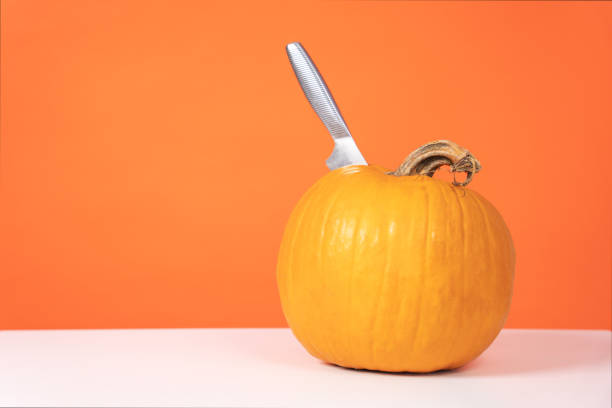 istock Ripe pumpkin with sharp knife sticking out of it, standing on the table. Preparing to create jack-o-lantern for decorating apartment for Halloween party, front view, copy space. Seasonal dishes. 1337523507