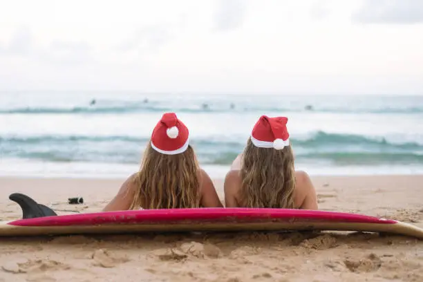 Two young adult women in bikinis and Santa hats relax on the beach after surfing on Christmas in Australia.