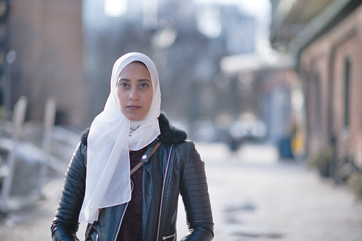 A young Muslim woman wearing a hijab standing outside on a street on a cold winter day. She is dressed warmly. She is looking at the camera with a serious expression on her face.