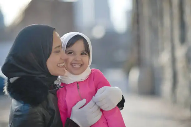 A beautiful Muslim mother wearing a hijab is giving her young daughter a hug. They are standing on a street outside on a winter day. She is smiling ear to ear at her daughter.