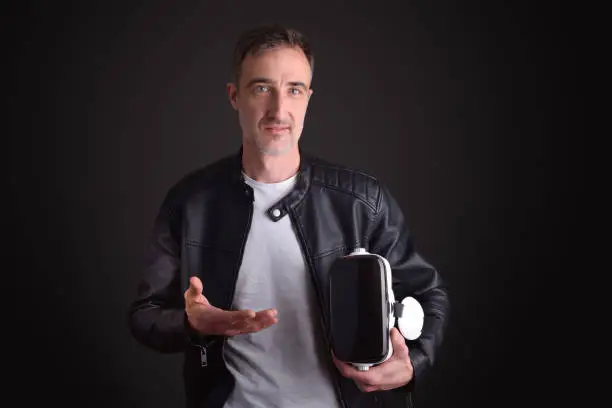 Man showing virtual reality headset dressed in white t-shirt and black leather jacket on black isolated background