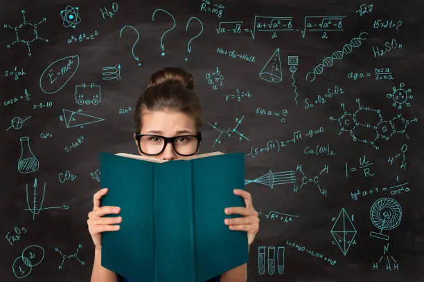 Photo of Student reading Open Book hiding Face. Shocked Young Girl in Eyeglasses behind Empty Blue Book Cover over Chalkboard with Math Formulas
