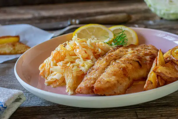 Delicious fish dish with fried redfish fillet served with chips or potato wedges and fresh coleslaw salad on a plate on rustic and wooden table background. Closeup and isolated view