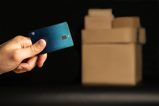 Hand with credit card, in front a black background. In the background not in focus cardboard boxes.