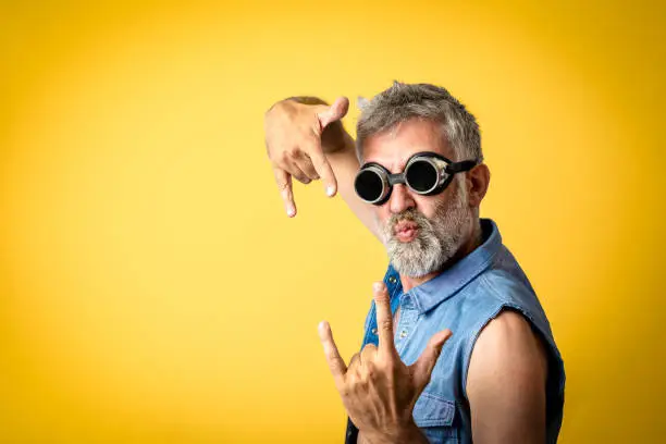 Photo of middle-aged man with beard wearing welding glasses on yellow background, funny facial expression of a male adult dressed as a worker metalmechanic making gesture of I love you with hands