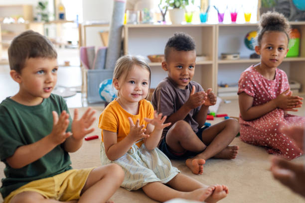 gruppe von kleinen kindergartenkindern, die drinnen im klassenzimmer auf dem boden sitzen und klatschen. - vorschulkind stock-fotos und bilder