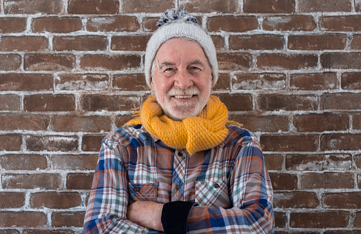 Portrait of attractive senior man wearing winter dress standing against a brick wall looking at camera smiling. Brick wall in background