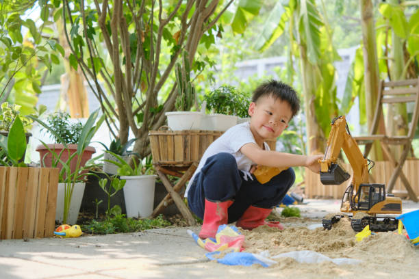 asiatisches lächelndes kind, das mit sandspielzeug und spielzeugbaumaschinen im garten des hinterhofs spielt - sandbox child human hand sand stock-fotos und bilder