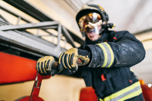 Anonymous firefighter in uniform with yellow lines and protective helmet moving red lever arm near ladder on fire truck in garage. Fire extinguishing concept