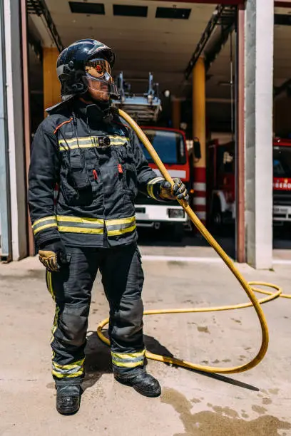 Photo of Anonymous firefighter in protective uniform with yellow lines standing with long hose dripping water on pavement in daylight