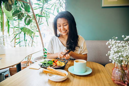 Young Asian woman with long loose black hair sits on sofa at wooden table and eats vegetable dish smiling in cosy vegan cafe closeup
