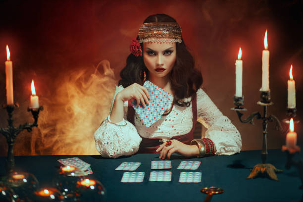 hermosa chica de fantasía en imagen de bruja gitana se sienta a la mesa en la habitación gótica oscura. traje rojo arte. adivina mujer mágica leyendo futuro en cartas del tarot. velas rituales encendidas, sesión de espiritismo, humo. - fortune teller fotografías e imágenes de stock