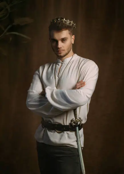 Portrait of handsome man in image of medieval king. Vintage clothing, historic white shirt retro clothing gold crown on head. Gothic room, studio. Fashion model posing. Adult strong sexy guy knight