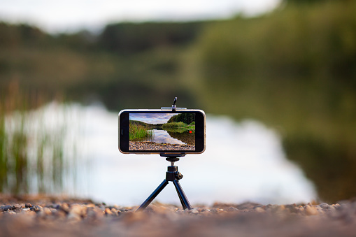 A close up of the phone on a tripod takes a video or a photo of nature. A beautiful lake in the forest with clouds in the screen of the photographer's mobile phone.
