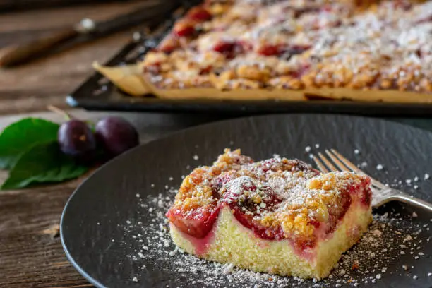 A piece of fresh baked plum crumble cake on a dark plate on rustic and wooden table. Whole cake is in the background. Closeup view