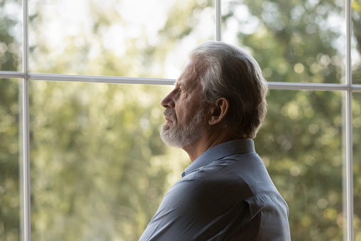 Pensive sad old Caucasian 60s man look in window distance feel lonely abandoned lack communication in retirement house. Unhappy thoughtful mature middle-aged male mourn grieve. Solitude concept.