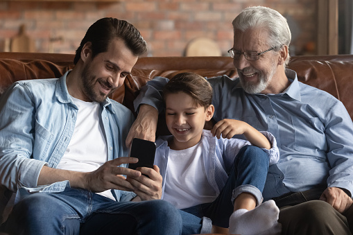 Smiling three generations of men sit relax on sofa in living room use cellphone gadget. Happy little Caucasian boy child with father and grandfather have fun talk on smartphone video webcam call.