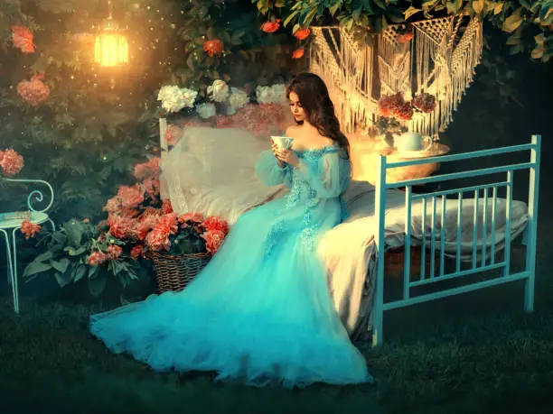 Photo of Artwork Fairy tale princess sits on vintage bed. Fantasy woman holding cup in hands drinking tea. Long wave hair. Blue vintage luxury dress. Backdrop blooming flowers, old lamp. Fantasy woman queen.