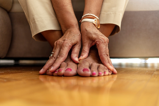 Close up of senior woman hands touching legs with varicose veins, sitting on sofa at home. Woman suffering from ankle pain. An older woman massages her feet to relieve the pain caused by arthritis.