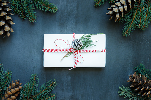 Christmas background with white wrapped gift box with red ribbon, sprig of conifer - juniper, pine cones, branches of spruce/fir on black stone background. Flat lay. New Year concept