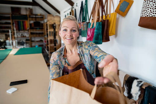 Personal perspective of a tanner passing a brown paper bag to a customer in her tannery studio after the customer has just bought a handmade leather bag from her. The business is committed to a zero waste approach and all the fabrics used are ethically sourced. / Female Focus Collection