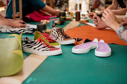 A group of unrecognisable women undertaking a leather shoe making workshop together. The main focus is finished custom designed trainers on the workbench and a pair of trainer soles. All the material used is ethically sourced and the business is committed to a zero waste approach. / Female Focus Collection