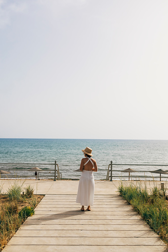 An unrecognizable young woman in a white dress standing on the dock, admiring the sea