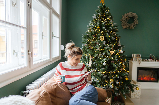 Photo of a young woman spending December day at home, drinking coffee and using smart phone. Christmas tree and holiday decoration in the background.