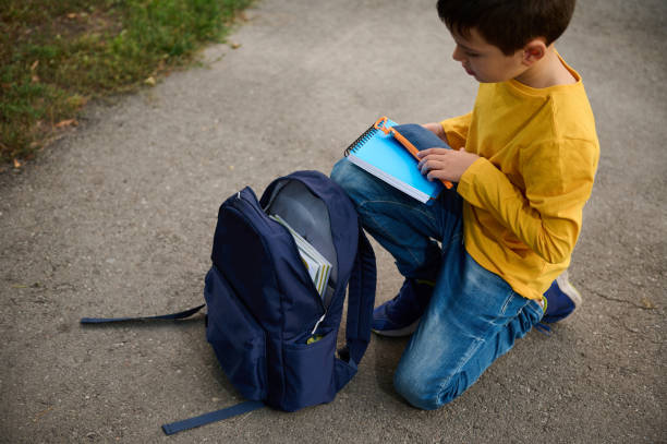 istock Charming elementary aged schoolboy sits on his knees on a path in the park, puts a notebook and pencil case in a backpack, returning home after shooting 1337646874
