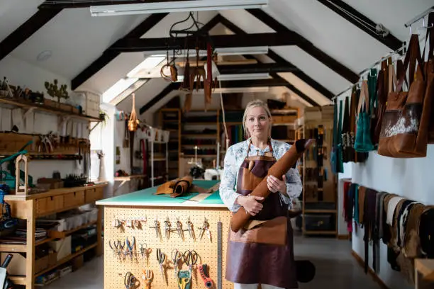 A three-quarter-length portrait view of an independent female small business owner. She is looking towards the camera with a proud look on her face as she stands in her Tannery leather shop and business workspace in Wales. / Female Focus Collection